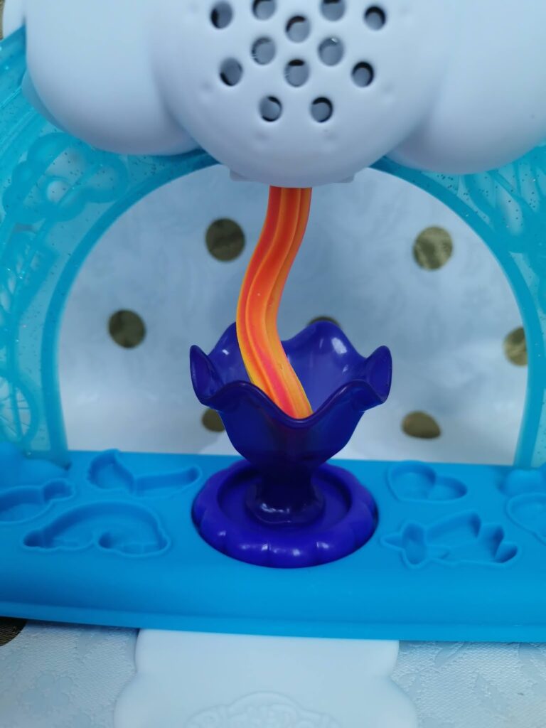 A close-up of a toy featuring a blue and purple flower-shaped bowl. Above it, a twisted orange and yellow ribbon or stream descends from a white fitting with small holes. The background is blue plastic with embossed shapes and white patterned fabric.