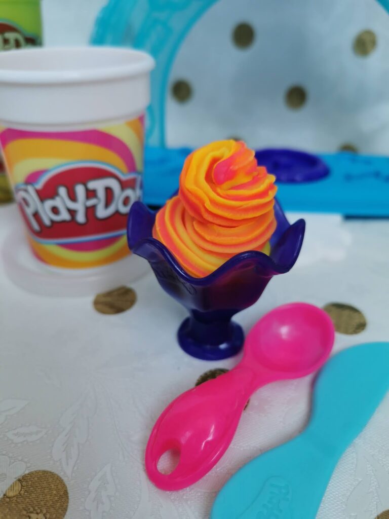 A purple toy dish holds orange and yellow swirled Play-Doh shaped like soft-serve ice cream. Nearby are a pink spoon and a blue spatula. In the background, there is a white Play-Doh container with a colourful label and a white surface with gold dots.