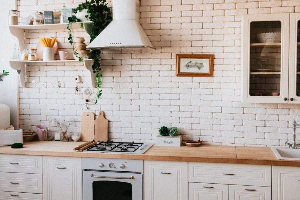 A bright kitchen with white brick walls, white cupboards, a wooden worktop, a gas hob, and an oven. Open shelves display plants, mugs, and jars. A white cooker hood is above the hob, and a glass cupboard and framed art are on the wall.