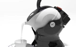 A futuristic, black and white electric kettle shaped like a sculpted helmet pours milk or hot liquid into a white teacup on a saucer. The design is modern and glossy, contrasting with the plain, bright background.