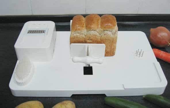 A white multipurpose kitchen chopping board features a loaf of bread secured by a handle, an integrated grater, a brush, and prongs for holding food. Surrounding the board are vegetables, including potatoes, a carrot, a cucumber, and an onion.