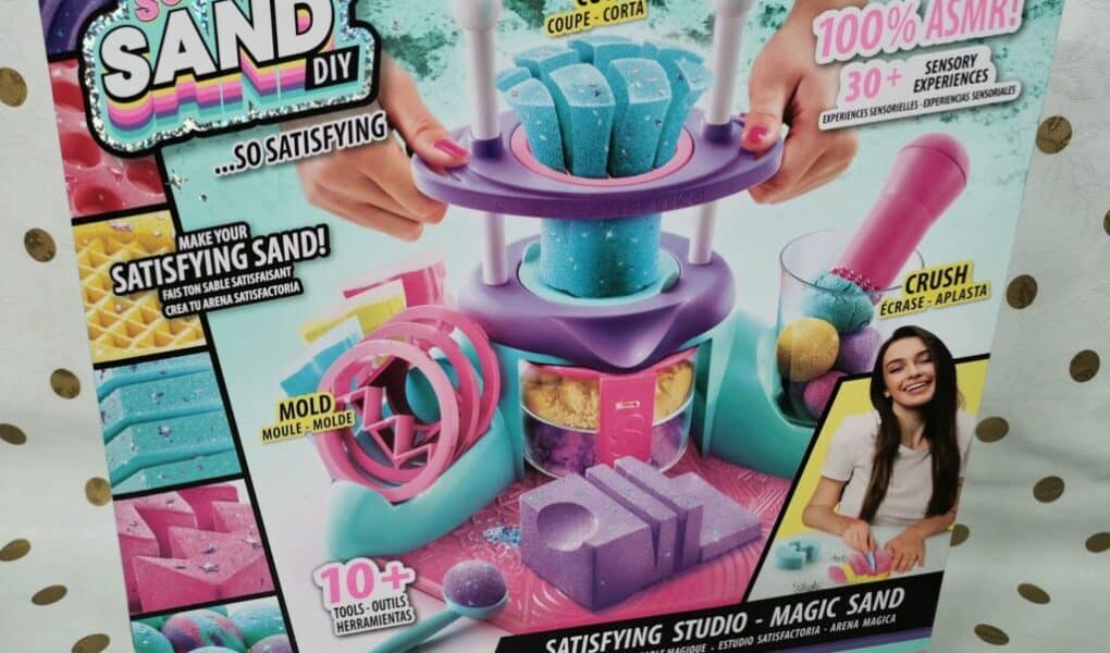 Colourful toy kit box labelled "So Sand DIY Satisfying Studio – Magic Sand." It shows a sand-moulding playset with purple, blue, and pink tools, moulds, and sand. Includes slogans like "100% ASMR!" and "Create shapes!" A smiling girl is pictured in the bottom right.