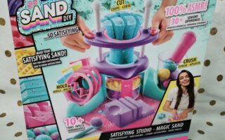 Colourful toy kit box labelled "So Sand DIY Satisfying Studio – Magic Sand." It shows a sand-moulding playset with purple, blue, and pink tools, moulds, and sand. Includes slogans like "100% ASMR!" and "Create shapes!" A smiling girl is pictured in the bottom right.