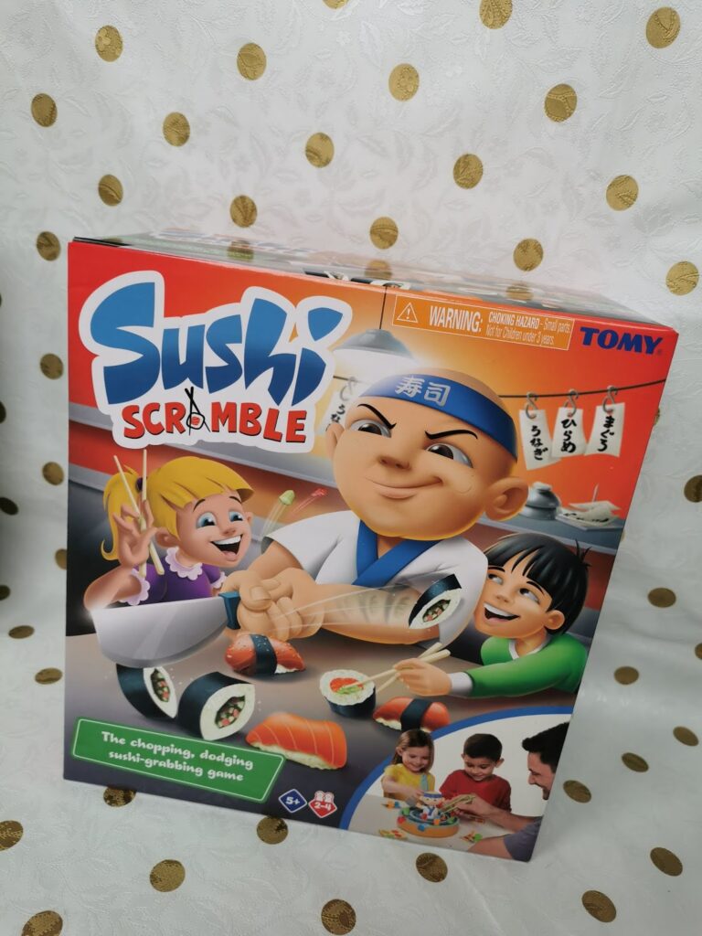 A colourful box for the game “Sushi Scramble” by Tomy is shown. The cover features animated characters: a smiling sushi chef and two excited children reaching for sushi. Bright sushi rolls and game pieces are depicted. Text reads: “The chopping, dodging sushi-grabbing game.”.
