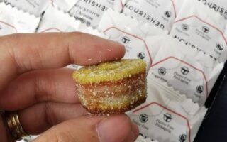 A hand holds a small, round, sugar-coated, layered gummy sweet with yellow and brown layers. In the background, several white packets labelled “NOURISHED” and “Plastic Free Packaging” are stacked neatly in a display box.