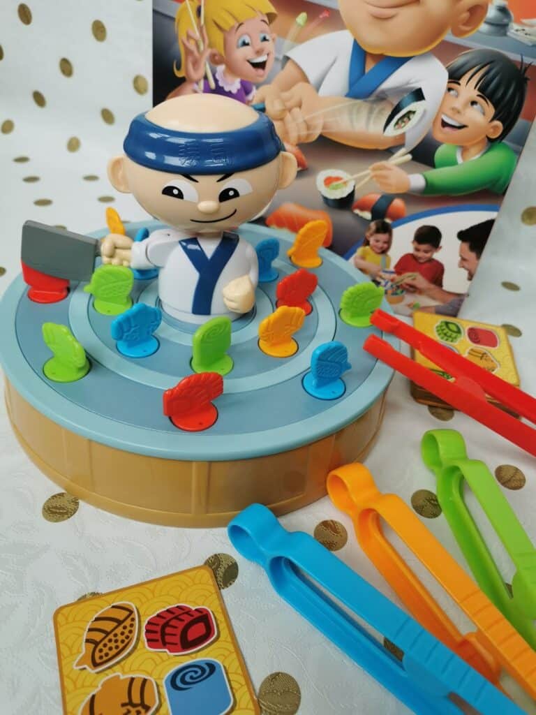 A sushi-themed children's game features a smiling chef figure at the centre atop a rotating tray with colourful sushi pieces. Surrounding the game are multicoloured tweezers and a game card, with the game's illustrated box in the background.