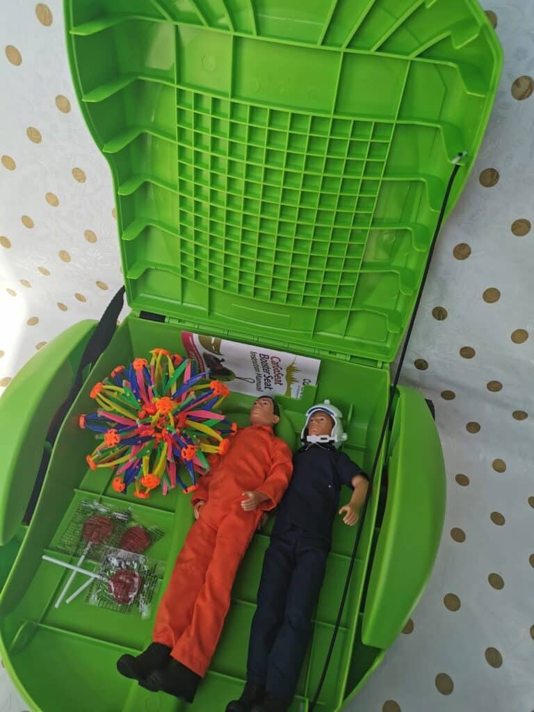 Two action figures—one in an orange suit, one in a blue suit with a white helmet—lie inside an open green plastic case. Nearby are four lollies, a colourful expandable ball toy, and a folded leaflet. The background is white with gold polka dots.