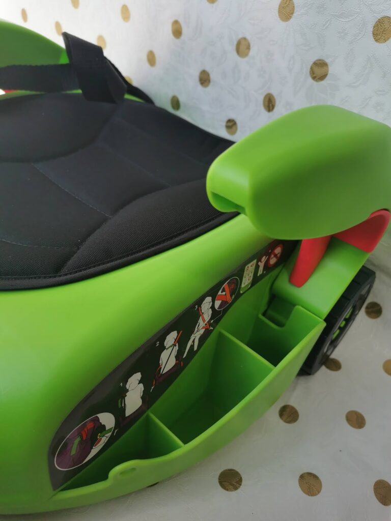 Close-up of a bright green child's ride-on toy car with a black seat and storage compartments. Safety instruction stickers with diagrams and red warnings are visible on the side. The background is white with gold spots.