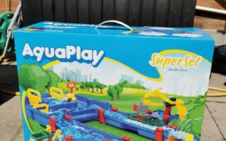 A large AquaPlay Superset box sits on a patio. The box shows a colourful toy water canal set with boats and accessories. The background includes a black plastic storage box, hose, and brick wall. The set is for ages 3–7, model number 1520.