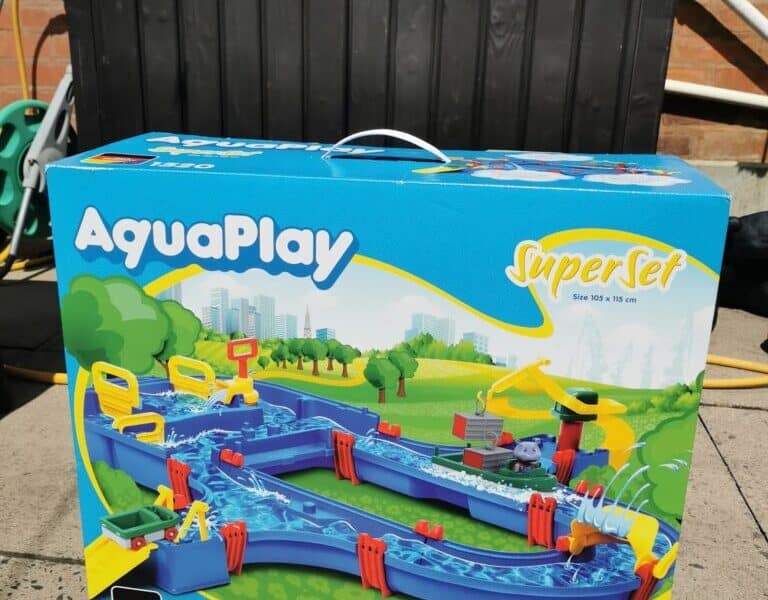 A large AquaPlay Superset box sits on a patio. The box shows a colourful toy water canal set with boats and accessories. The background includes a black plastic storage box, hose, and brick wall. The set is for ages 3–7, model number 1520.