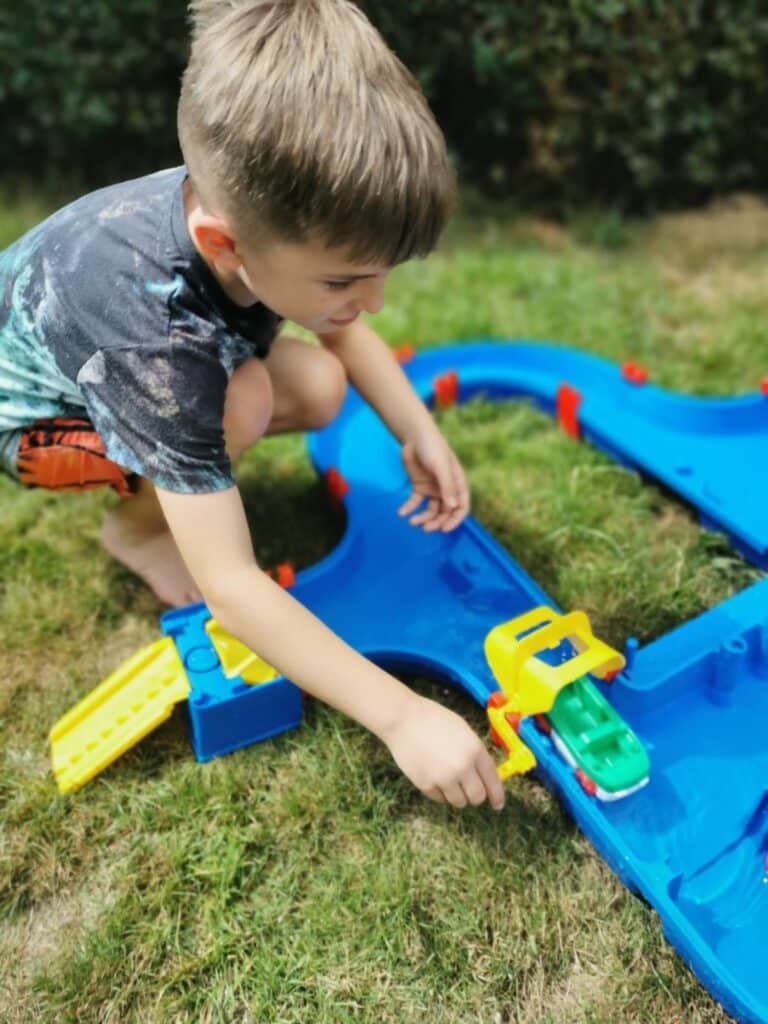 A young boy with light brown hair and a black-and-grey shirt kneels on grass, playing with a blue plastic waterway toy. He guides a green and yellow toy boat along the water track, appearing focused and engaged outdoors.