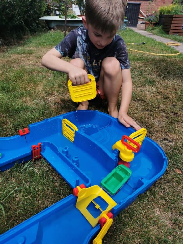 A young boy kneels on grass, playing with a blue plastic waterway toy. He pours water with a yellow container into the canal, moving toy boats and gates. The scene is outdoors in a garden with plants, bricks, and a yellow hose in the background.