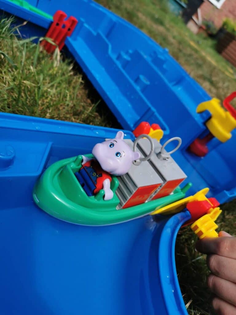 A toy hippo figure in a green boat floats in a blue plastic waterway playset on grass. The boat has a red and grey cargo section, and a child’s hand is visible turning a yellow and red crank on the playset. The scene is outdoors.