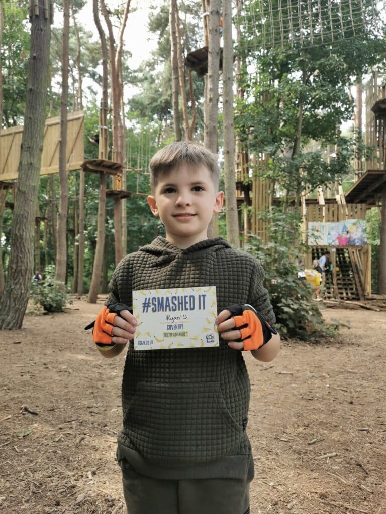 A young boy stands in a forest adventure park, wearing a green hoodie and orange gloves. He is smiling and holding a sign that reads "#SMASHED IT, Saying I’ve completed CONFIDENCE." Wooden platforms and ropes are visible among tall trees in the background.