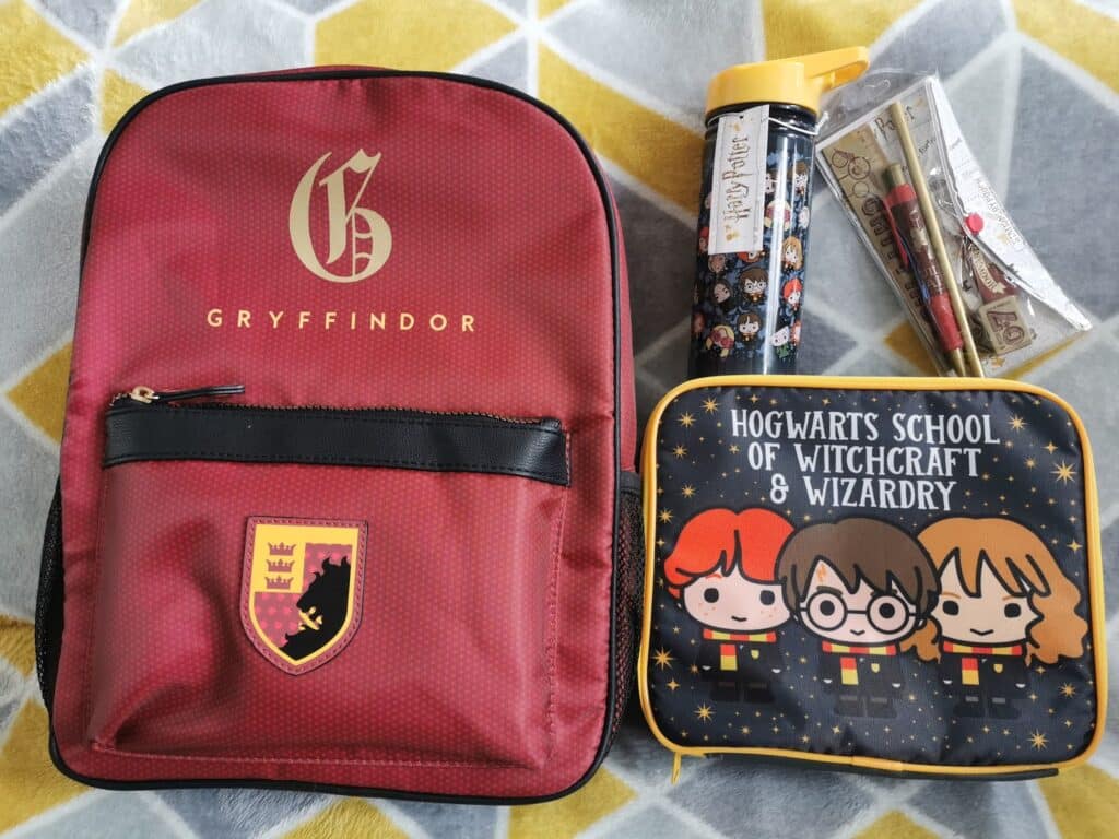 A maroon Gryffindor rucksack with gold lettering and crest, a Hogwarts lunch bag with cartoon Harry, Ron, and Hermione, a Harry Potter-themed water bottle, and a clear pencil case with Harry Potter graphics are placed on a grey and yellow patterned blanket.