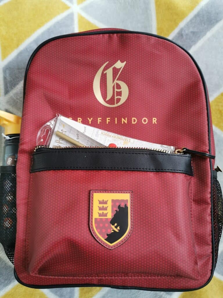A red Gryffindor-themed rucksack with a gold "G" and "GRYFFINDOR" text on top. The front pocket is partly unzipped, revealing papers inside. The Gryffindor crest patch is below the pocket. The rucksack rests on a grey and yellow patterned surface.