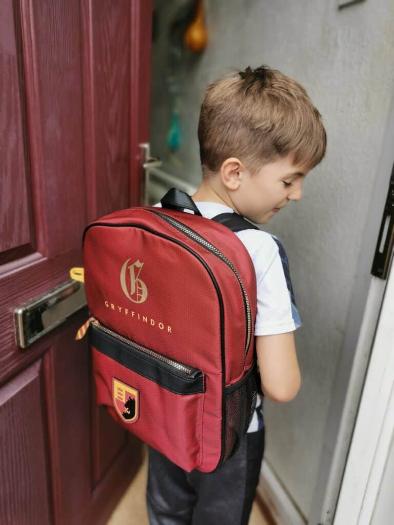 A young boy with light brown hair faces sideways, wearing a red Gryffindor rucksack with gold lettering and a crest. He stands near an open maroon door, ready to step outside, with part of a letterbox and a grey wall visible.