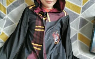 A child wearing a black Hogwarts robe with maroon and yellow stripes, and a hood partially covering their face, sits on a grey and yellow geometric patterned blanket. The Gryffindor crest is visible on the robe, and the child smiles softly.