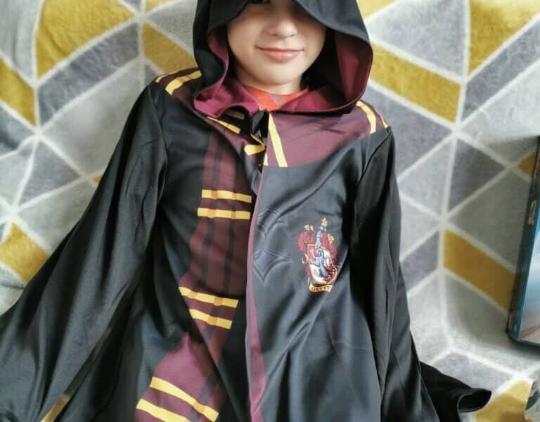 A child wearing a black Hogwarts robe with maroon and yellow stripes, and a hood partially covering their face, sits on a grey and yellow geometric patterned blanket. The Gryffindor crest is visible on the robe, and the child smiles softly.