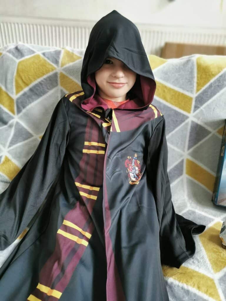 A child wearing a black hooded robe with maroon and gold stripes, featuring a Hogwarts crest, sits on a sofa with a geometric-patterned blanket. The child is smiling, with the hood partially covering their face.
