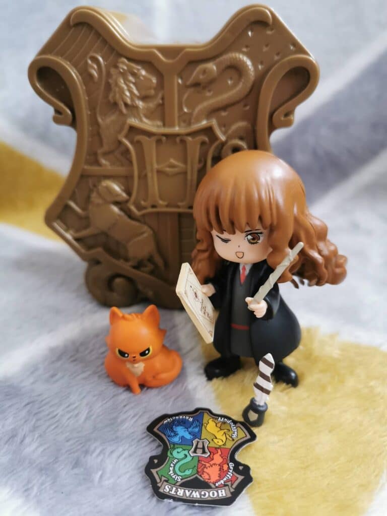 A Hermione Granger figurine in Hogwarts robes holds a wand and book. Beside her is a small orange Crookshanks figure. Behind them is a brown Hogwarts crest with animal emblems. In front is a colourful "Hogwarts" crest badge on a soft surface.