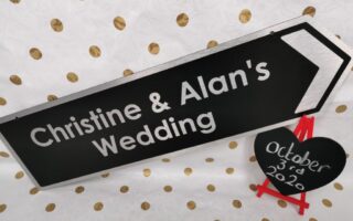 A black sign with white text reads, "Christine & Alan's Wedding," with a white arrow pointing right. Next to it, a small black heart-shaped chalkboard on a red easel displays "3rd October 2020." The background is white with gold polka dots.