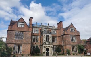 A large historic red-brick manor with tall chimneys, many windows, and ornate stonework above the main entrance. The building is surrounded by trimmed shrubs, a gravel path, and a manicured lawn under a partly cloudy blue sky.