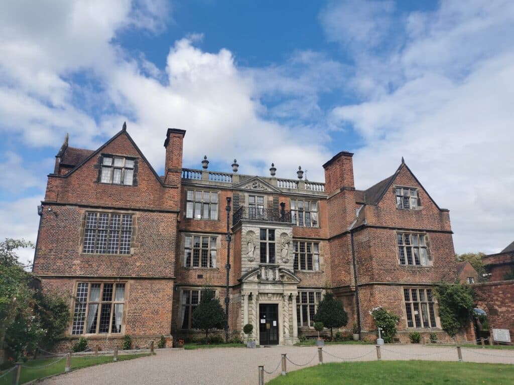 A large historic brick manor with tall chimneys, ornate stone entrance, and many multi-paned windows. Two symmetrical wings flank the central section. The sky is partly cloudy, and the building is surrounded by greenery and a gravel path.