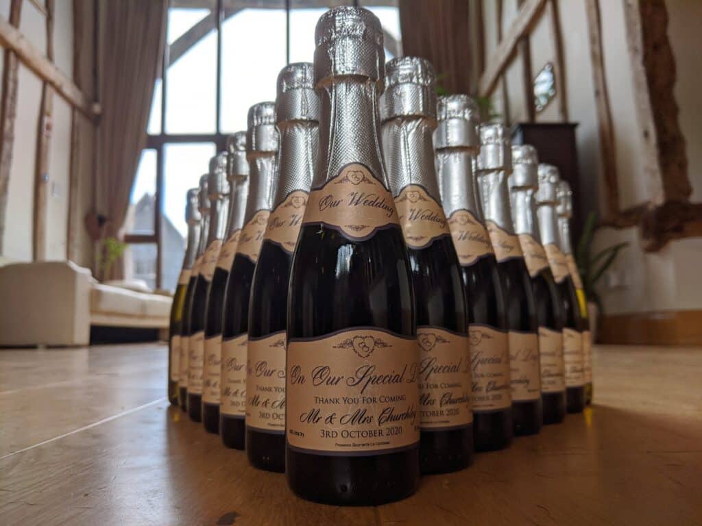 A group of small champagne bottles with cream-coloured labels reading “Our Special Day” are arranged in rows on a wooden floor. The background shows large windows, exposed wooden beams, and a cream sofa in a warmly lit, rustic room.