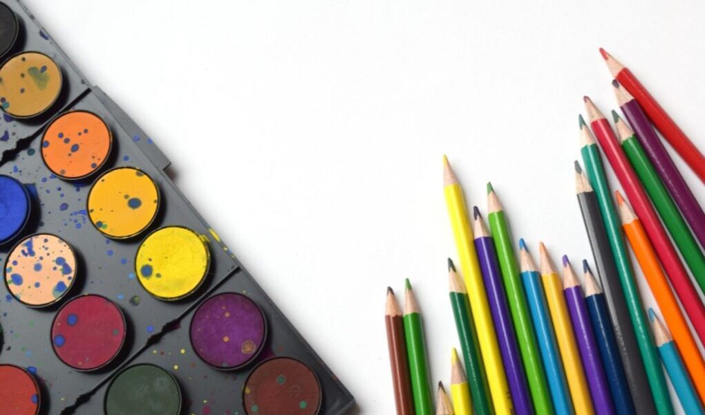 A set of watercolour paints with splatters sits on the left, showing various colours. On the right, coloured pencils in shades of yellow, green, blue, red, and purple are arranged with points facing upward on a white background.