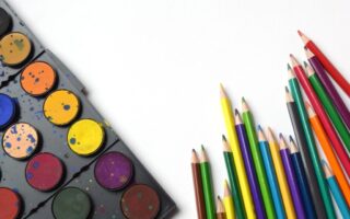 A set of watercolour paints with splatters sits on the left, showing various colours. On the right, coloured pencils in shades of yellow, green, blue, red, and purple are arranged with points facing upward on a white background.