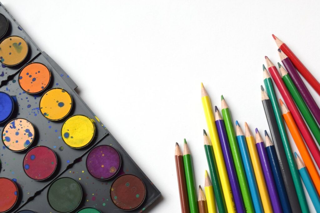 A watercolour palette with colourful paint circles and splatters rests on the left, while sharpened coloured pencils in various bright colours are arranged on the right, all against a white background.