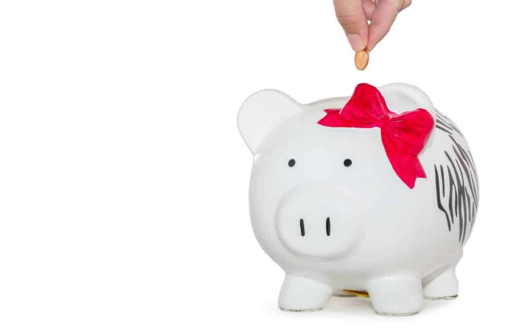 A hand drops a coin into a white piggy bank decorated with a large red bow. The background is plain white, emphasising the piggy bank and the coin. The image portrays saving money or financial planning.