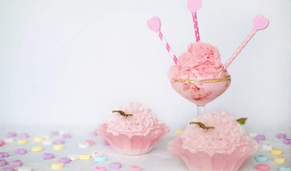 A glass with scoops of pink ice cream is decorated with three pink heart-topped sticks. In front, two pink cupcake-like desserts sit in cases. Colourful sweet hearts are scattered on a white surface, and the background is plain white.
