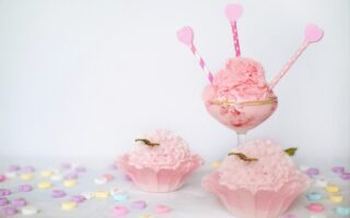 A glass with scoops of pink ice cream is decorated with three pink heart-topped sticks. In front, two pink cupcake-like desserts sit in cases. Colourful sweet hearts are scattered on a white surface, and the background is plain white.