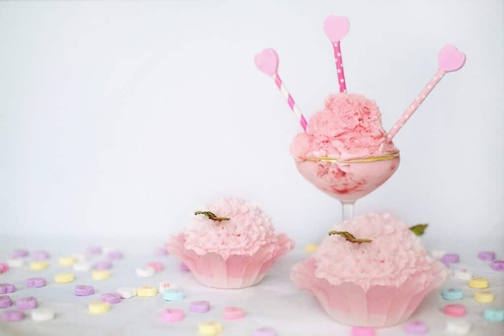 A glass dish filled with pink ice cream and topped with three pink heart-decorated sticks sits beside two pink frosted cupcakes in ruffled cases. Colourful pastel sweet hearts are scattered across the white surface. The background is plain white.