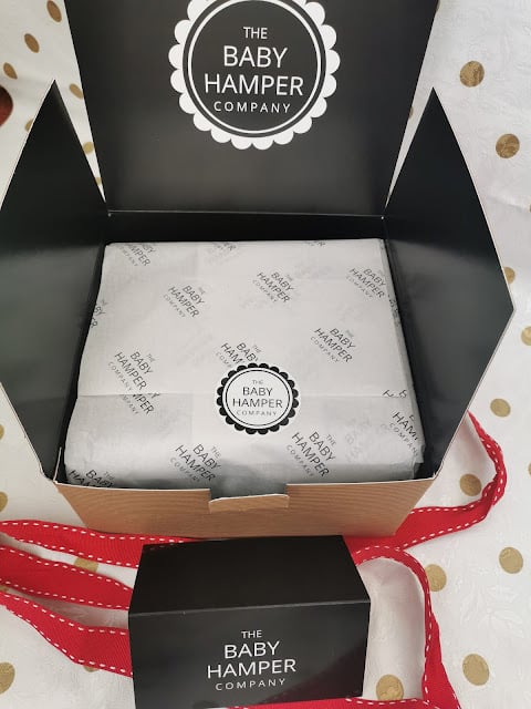 A cardboard gift box from The Baby Hamper Company sits open, revealing white tissue paper with the company logo printed in black. A black sticker with the logo seals the tissue. A matching smaller closed box and red ribbon are nearby.