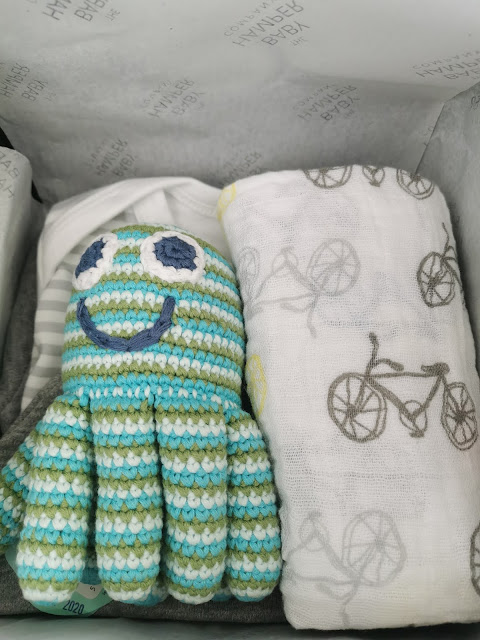 A crocheted octopus toy with blue, green, and white stripes and a smiling face sits beside a white cloth featuring a grey bicycle pattern, both placed in a box lined with tissue paper.
