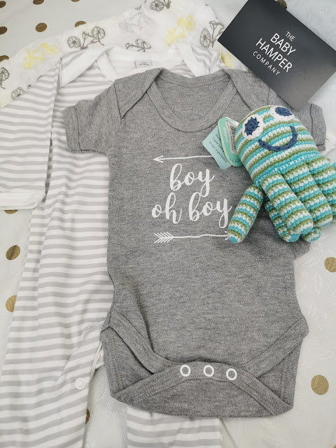 A grey baby babygrow with "boy oh boy" written on it is laid on top of a white and grey striped sleepsuit. Next to the babygrow is a blue and green crocheted toy, and a black card labelled "The Baby Hamper Company" is above them.