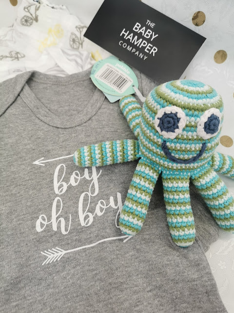 A grey baby onesie with “boy oh boy” written in white is next to a blue, green, and white striped crocheted octopus toy. Behind them is a black tag reading “The Baby Hamper Company.” White and yellow patterned fabric is partially visible.