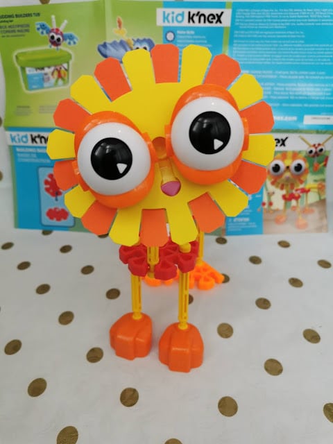 A colourful toy creature made from Kid K'Nex pieces stands on a white surface with gold polka dots. It has large, round eyes, orange and yellow parts forming a spiky face, red joints, and orange feet. Instructions and box are blurred in the background.