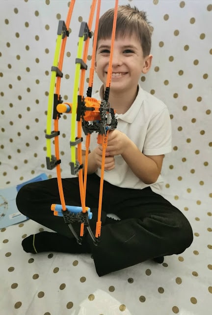 A young boy smiles whilst sitting cross-legged on the floor, holding a toy bow made of bright orange and yellow plastic pieces. He wears a white polo shirt and black trousers. The background is white with gold polka dots.