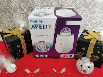 A Philips Avent bottle warmer box sits on a red surface, flanked by two black gift boxes with gold ribbons and white bird decorations. A glittery reindeer ornament is in front. The background features a grey-and-white patterned wall.