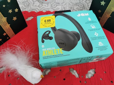 A teal box of JAM True Wireless Athlete earbuds sits on a red surface with a white feathered bird decoration nearby. The box features an image of the earbuds, product details, and a yellow sticker highlighting 6-hour battery life. Decorative items surround the box.