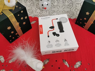 A white box of a Joby FreeHold smartphone grip lies on a red mat with a feathered bird decoration. The box features product images and instructions. Festive green gift boxes with gold ribbons and star patterns are placed nearby on a grey patterned surface.