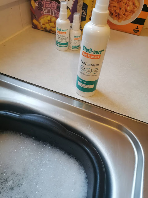 A bottle of Steri-eeze Hand Sanitiser sits on a kitchen worktop next to a sink filled with soapy water. Two more bottles and a cereal box are visible in the background on the cream-coloured worktop.