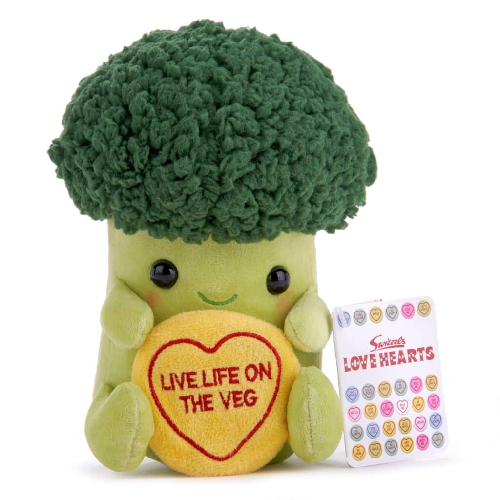 A plush broccoli toy with a smiling face holds a yellow heart-shaped cushion that reads "LIVE LIFE ON THE VEG." Next to it is a pack labelled "Swizzels LOVE HEARTS" featuring colourful sweet designs with messages.