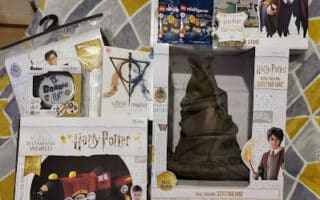 A collection of Harry Potter-themed merchandise displayed on a yellow quilt. Items include a Hogwarts Express draught excluder kit, a real-talking Sorting Hat, a Harry Potter board game, a Sorting Hat pin, a LEGO set, a wand kit, and platform 9¾-themed accessories.