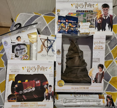 A collection of Harry Potter-themed merchandise displayed on a yellow quilt. Items include a Hogwarts Express draught excluder kit, a real-talking Sorting Hat, a Harry Potter board game, a Sorting Hat pin, a LEGO set, a wand kit, and platform 9¾-themed accessories.