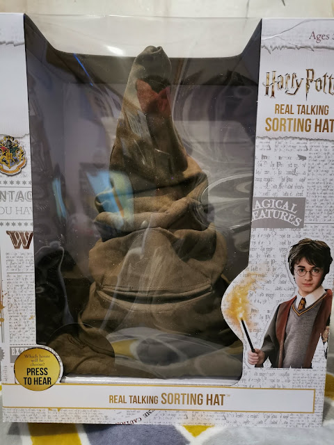 A boxed Harry Potter “Real Talking Sorting Hat” toy sits in its packaging. The brown, wrinkled hat with stitched facial features is visible through a clear plastic window. The box features Harry Potter graphics, with an image of Harry holding a wand and a “Press to Hear” button.